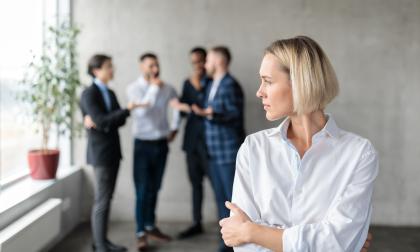 Male Coworkers Whispering Behind Back Of Unhappy Businesswoman Spreading Rumors And Gossips Standing In Modern Office. Sexism And Bullying Problem At Workplace Concept. Selective Focus