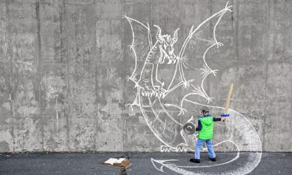 Little 5 year old boy fighting an imaginary dragon using a wooden sword and kitchen accessories as armor. Importance of reading and existence of non-visual media. Dragon chalk drawing on the concrete wall. Natural light.