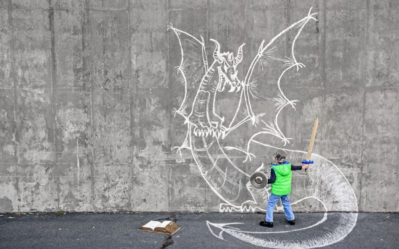 Little 5 year old boy fighting an imaginary dragon using a wooden sword and kitchen accessories as armor. Importance of reading and existence of non-visual media. Dragon chalk drawing on the concrete wall. Natural light.